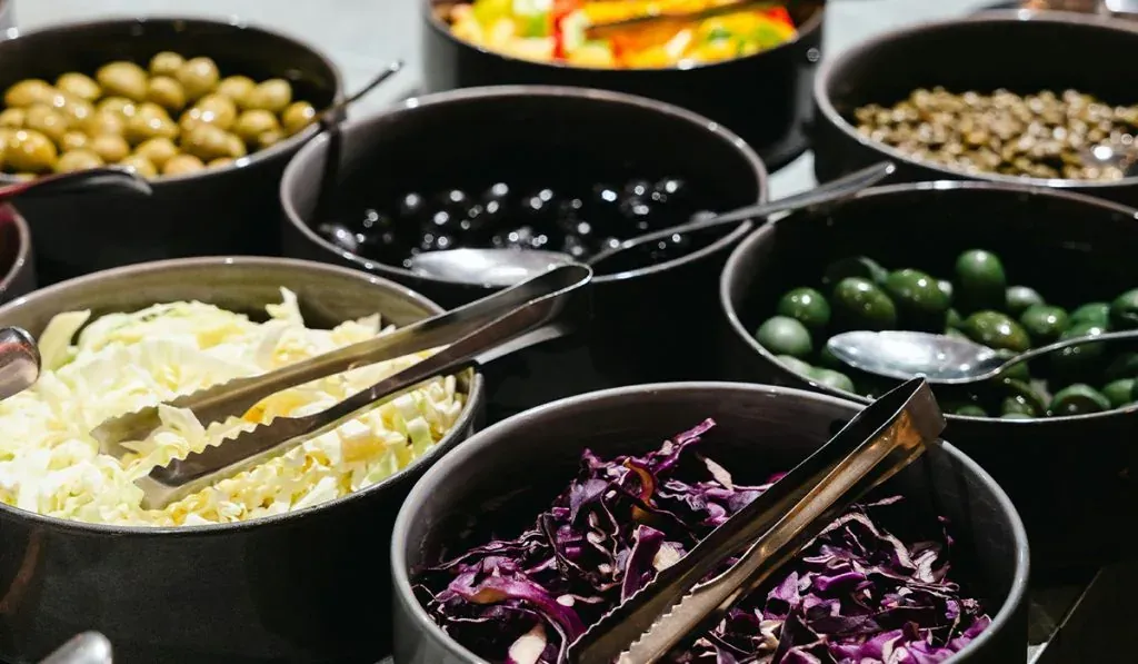Nahaufnahme eines Selbstbedienungsbuffets mit verschiedenen Zutaten in Schüsseln. Zu sehen sind grüne und schwarze Oliven, gehackter Weiß- und Rotkohl, sowie andere frische Zutaten. Jede Schüssel hat eine Zange oder einen Löffel zum Servieren.